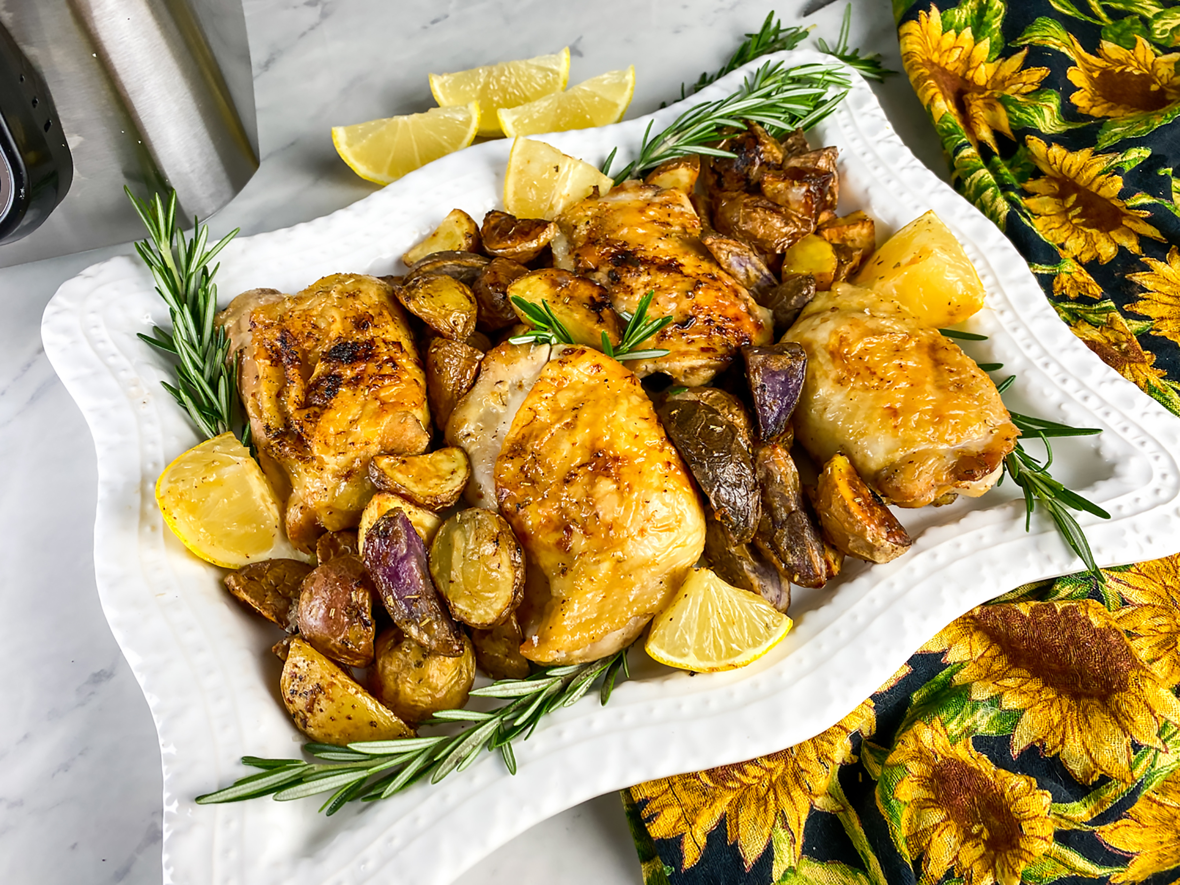 Roasted chicken thighs with crispy skin served on a white platter alongside golden baby potatoes, fresh rosemary sprigs, and lemon wedges, creating a flavorful and inviting dish perfect for a family meal.