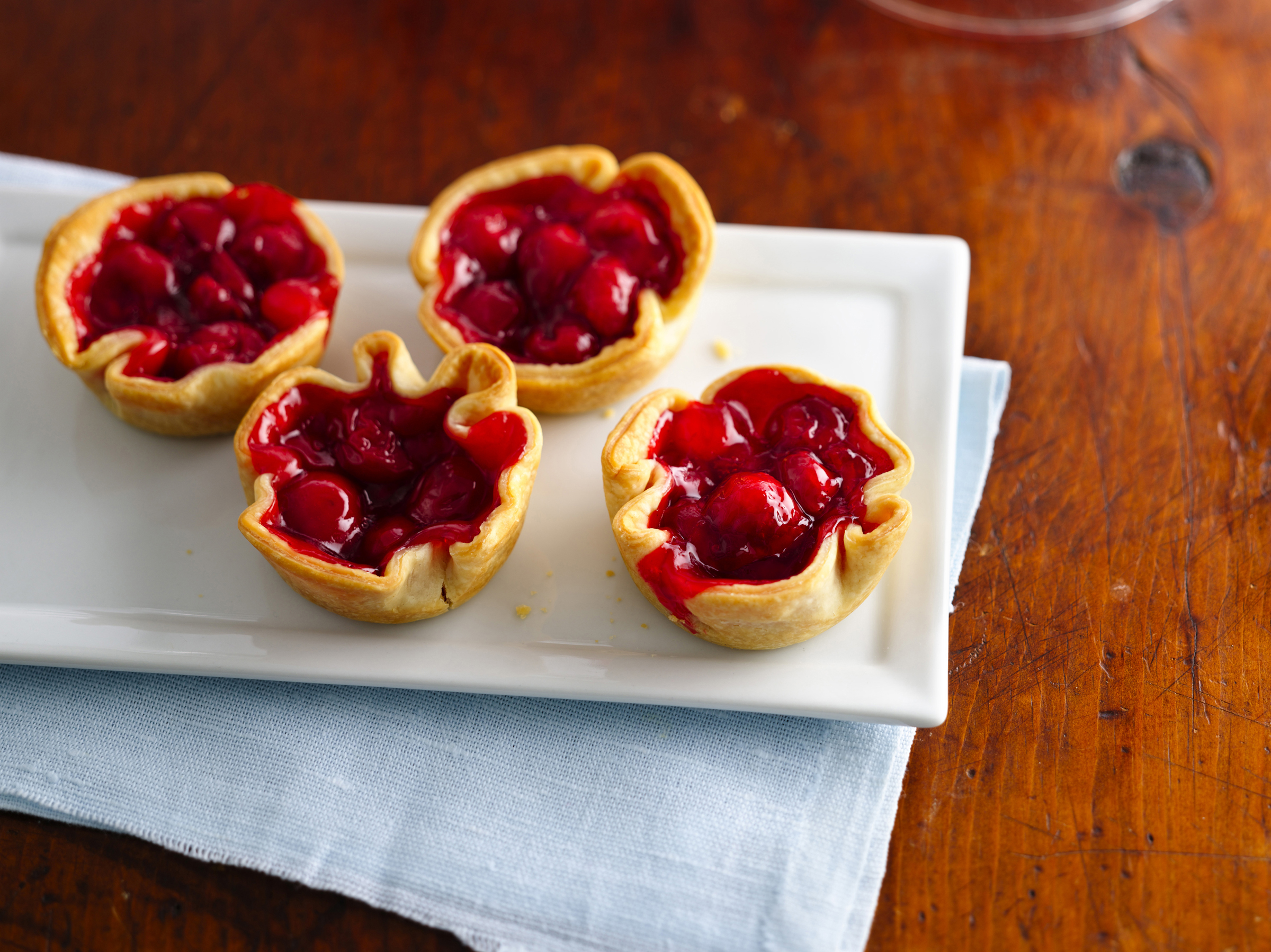 Cherry tartlets with a flaky crust, filled with glossy cherry filling, presented on a white plate over a light blue cloth, perfect for dessert lovers and special occasions.