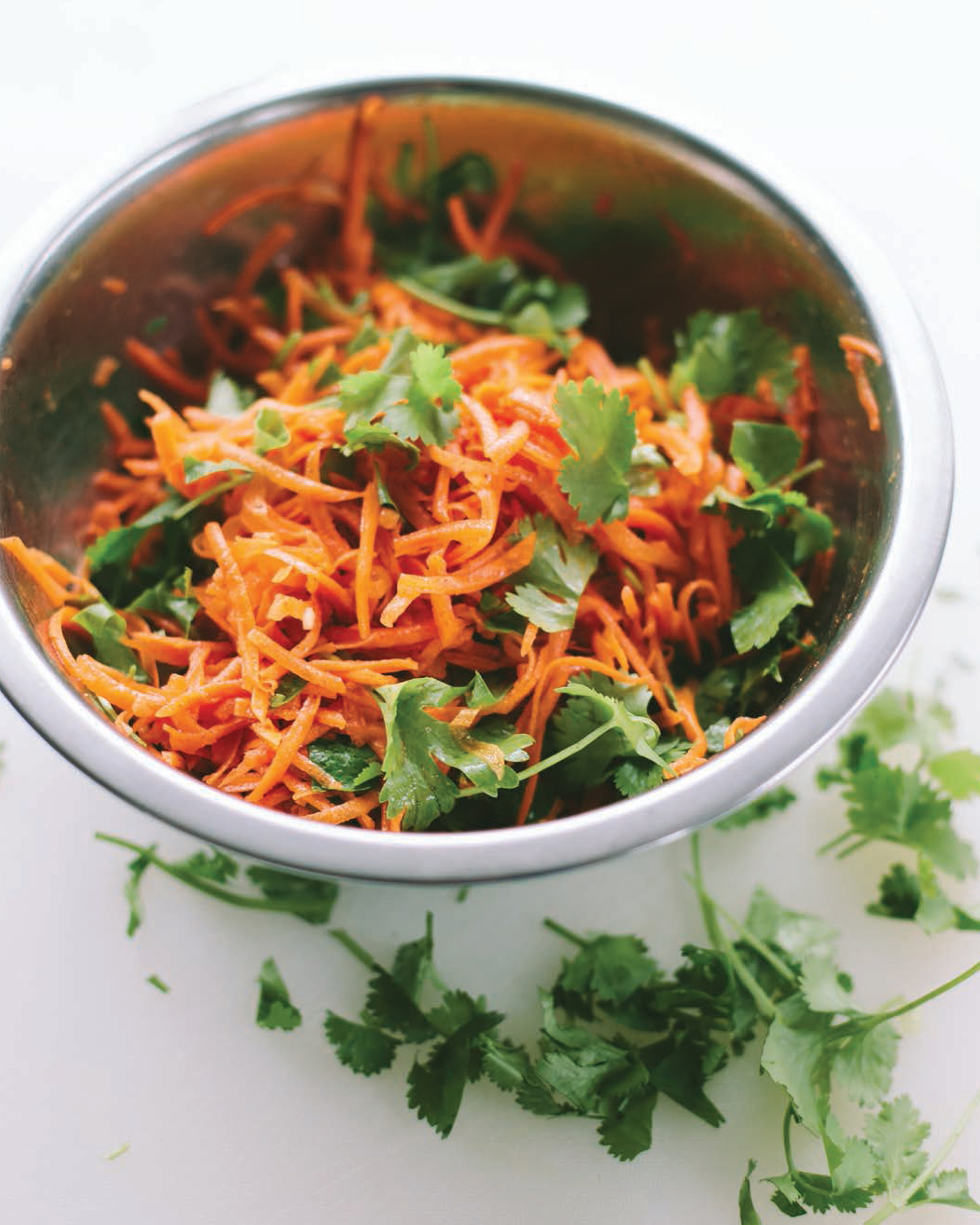 Freshly shredded carrots mixed with vibrant cilantro in a silver mixing bowl, surrounded by scattered cilantro leaves on a white surface, perfect for a healthy salad or garnish.
