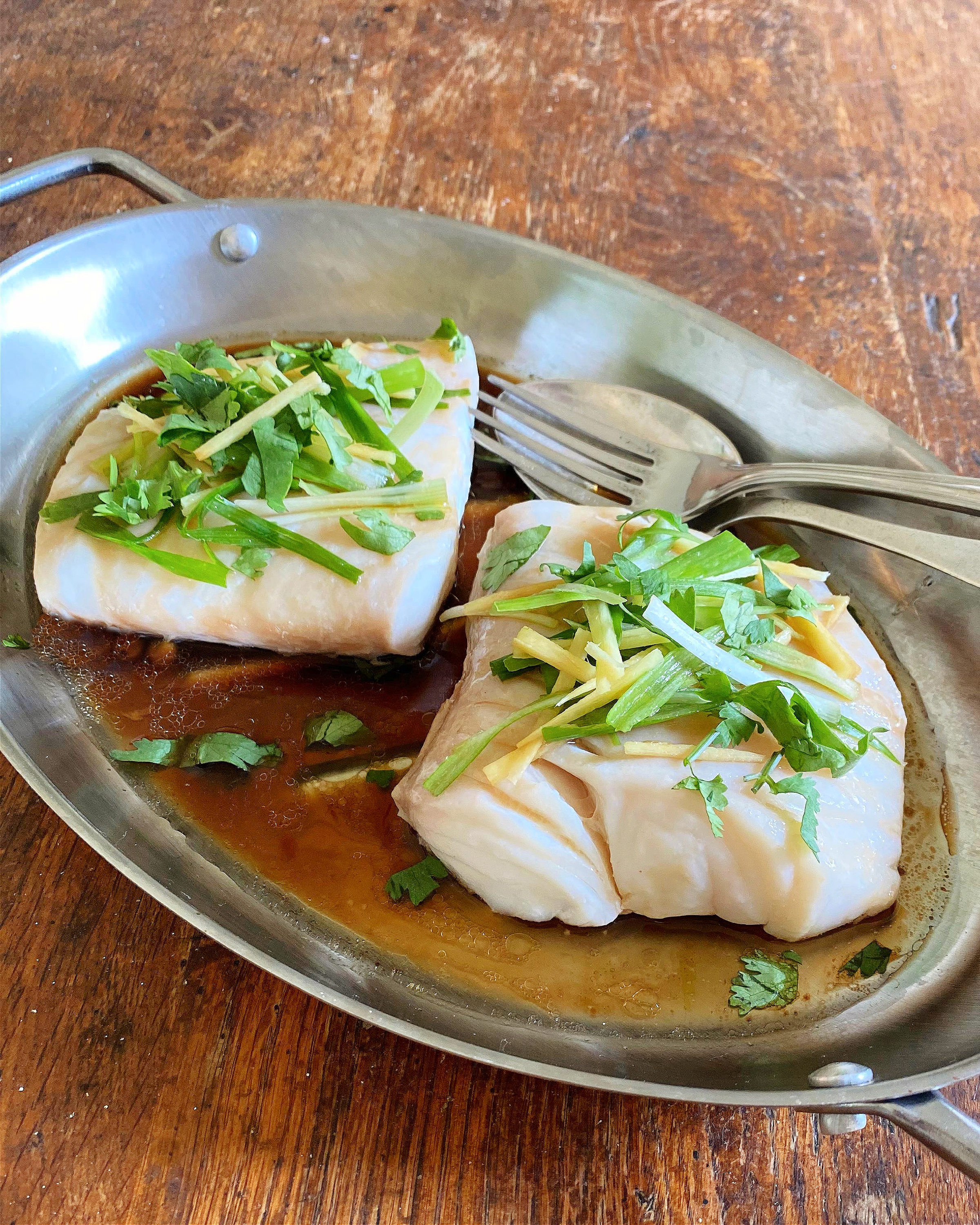 Steamed fish fillets garnished with fresh cilantro and ginger, served in a silver platter with a savory sauce, on a rustic wooden table. Perfect for healthy meal ideas and Asian-inspired recipes.