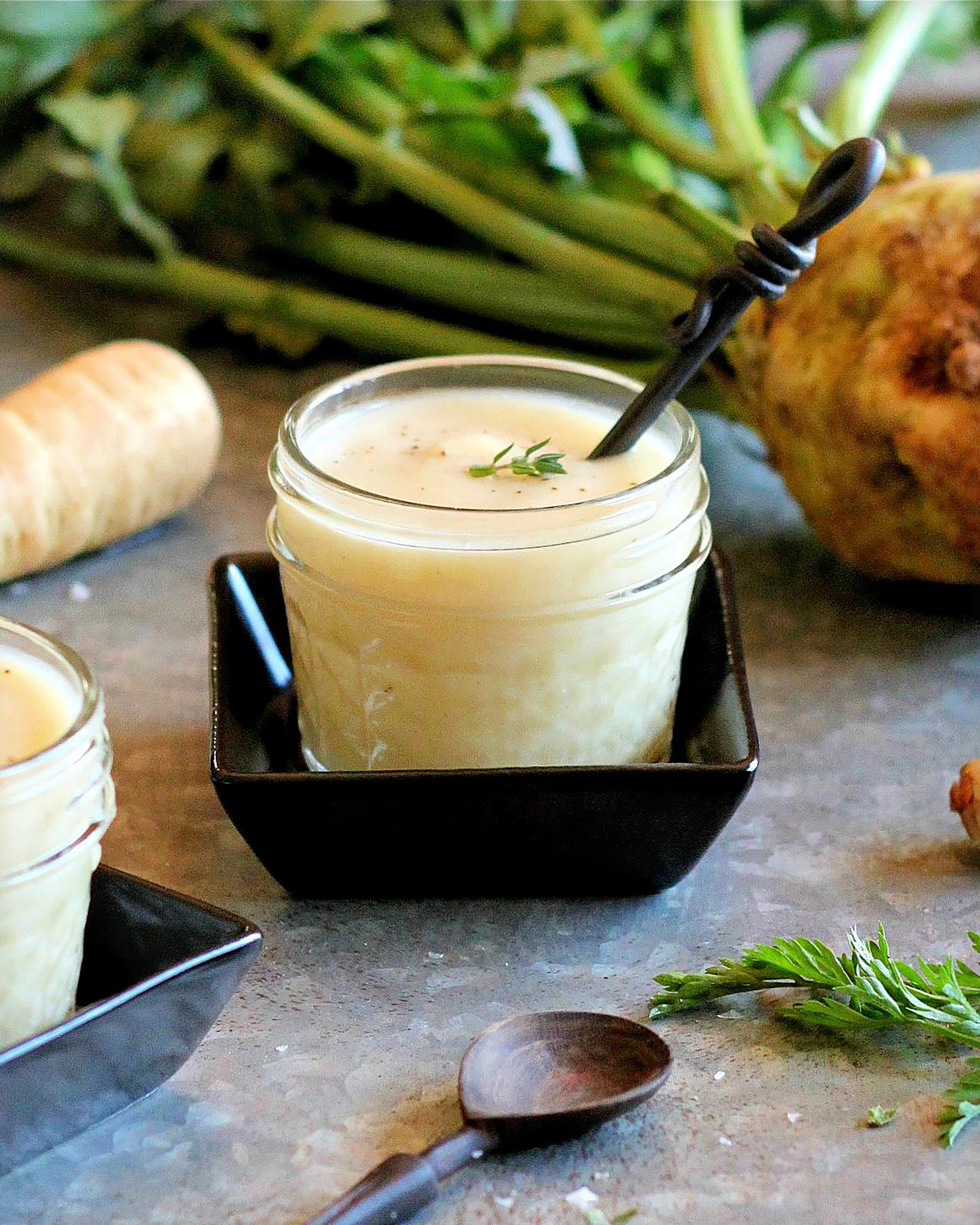 Creamy white soup served in a glass jar with a black spoon, garnished with fresh thyme, alongside parsnips and celery leaves on a textured surface. Ideal for a healthy and comforting meal.