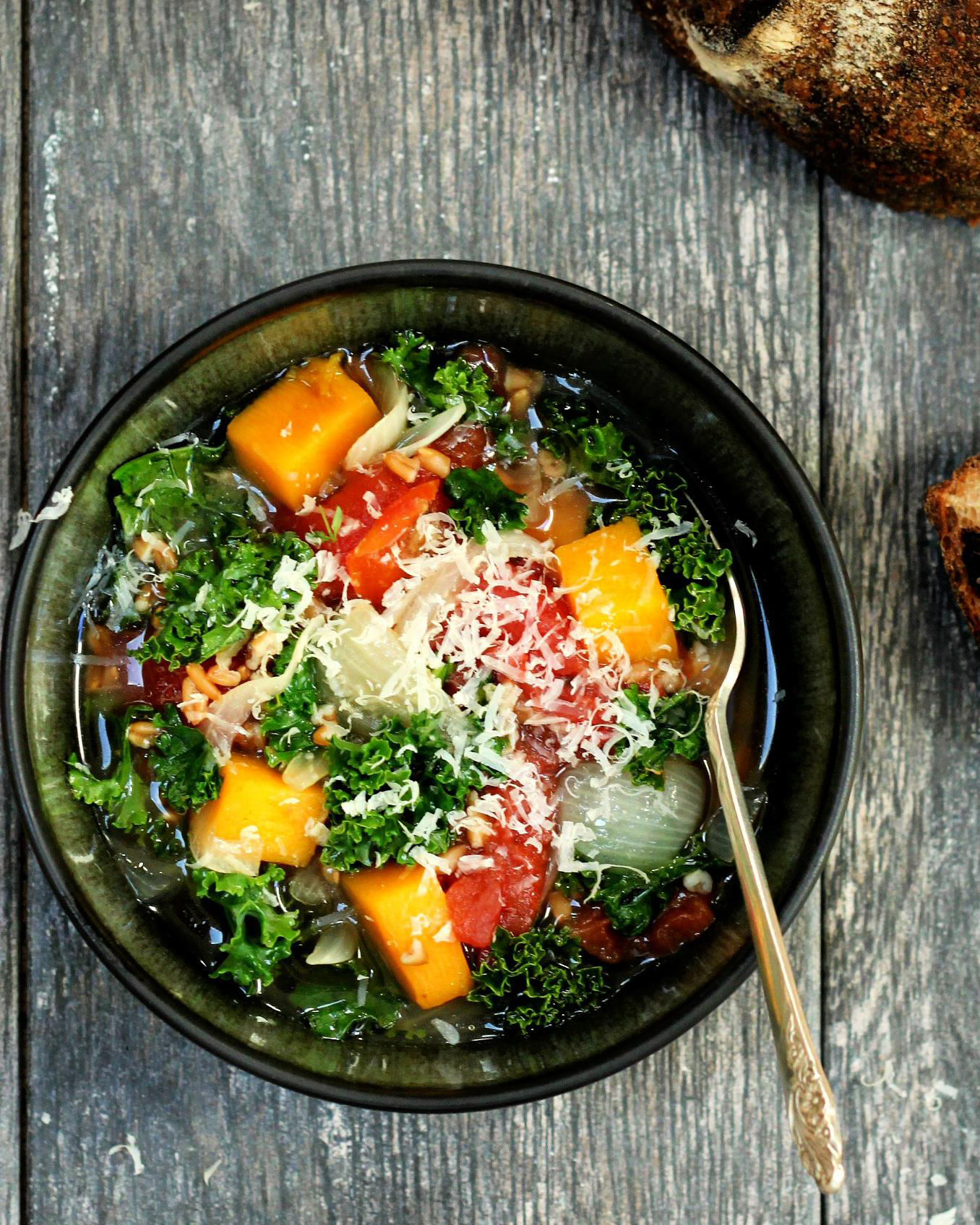 Colorful vegetable soup with kale, tomatoes, and butternut squash, topped with grated cheese, served in a dark bowl on a rustic wooden table alongside slices of crusty bread.