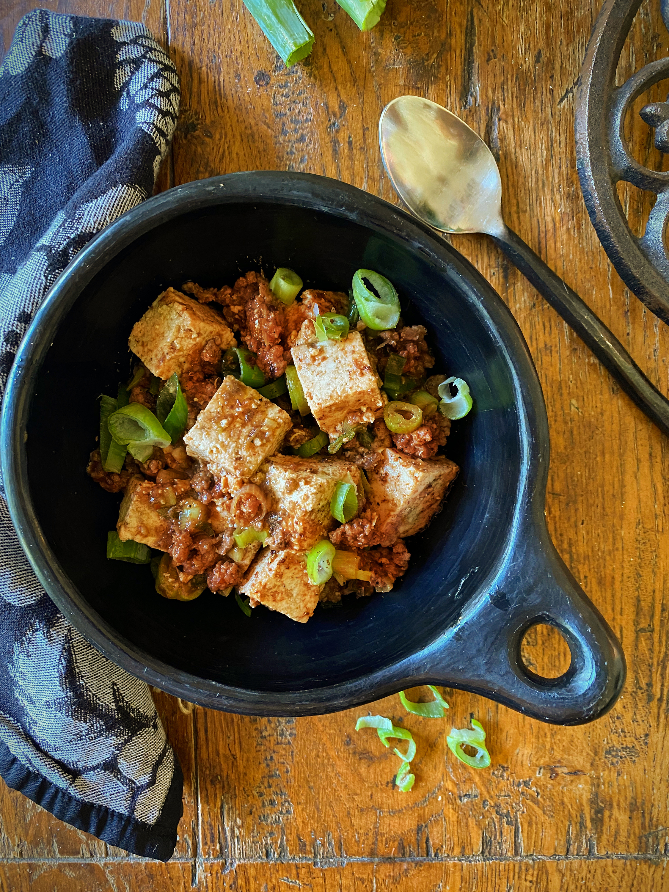 Bowl of tofu and ground meat stir-fry topped with green onions, served on a rustic wooden table with a spoon and a patterned napkin nearby.